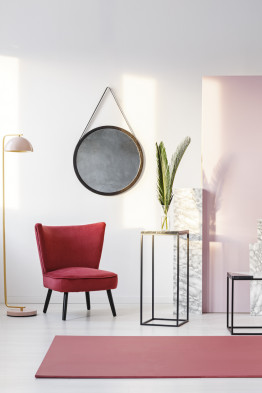 Round mirror hanging on white wall in room interior with burgundy velvet chair, red carpet and fresh leaves in glass vase